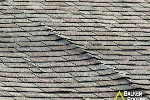Close-up of damaged asphalt roof shingles showing buckling and warping from possible water or heat damage.
