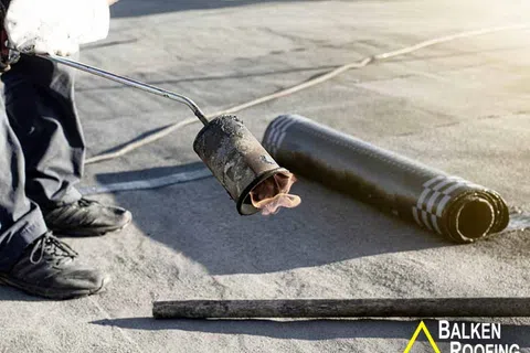 Worker using a torch to apply roll roofing material on a flat roof during installation process.