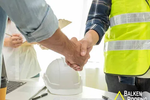 Two professionals shaking hands over roofing plans with a hard hat and laptop on the table.
