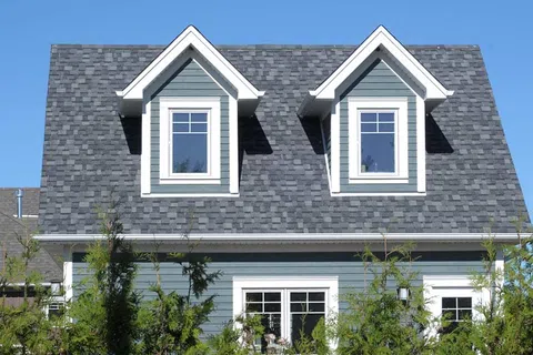 Modern blue house with gray shingled roof and white framed windows behind green shrubs under clear sky