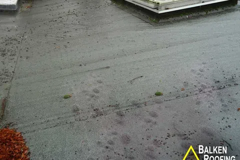 Flat rooftop with waterproofing membrane, drainage structures, and scattered leaves on surface.