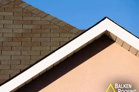 Close-up of brown shingle roof and beige wall under clear blue sky with Balken Roofing logo.