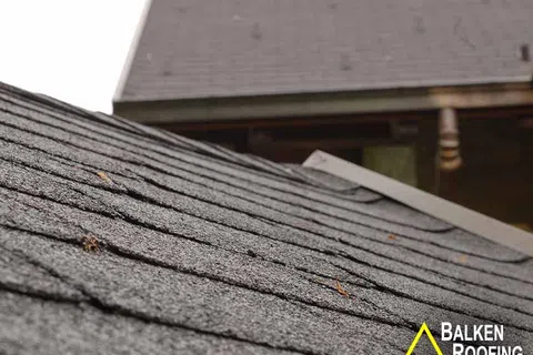 Close-up of textured gray asphalt shingle roof with a house background and Balken Roofing logo.