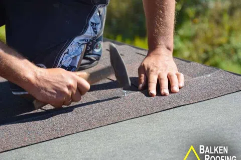 Close-up of hands hammering nails into asphalt shingles on a roof under sunny daylight.