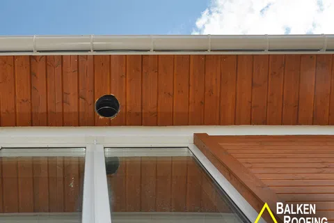 Wood-paneled roof soffit with a round vent hole above white-framed glass windows under blue sky.