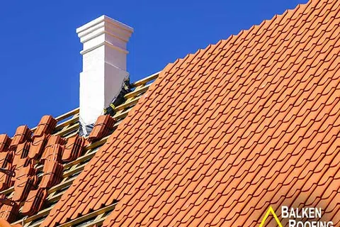 Roofers installing orange clay tiles on a sloped roof with a white chimney under a clear blue sky.