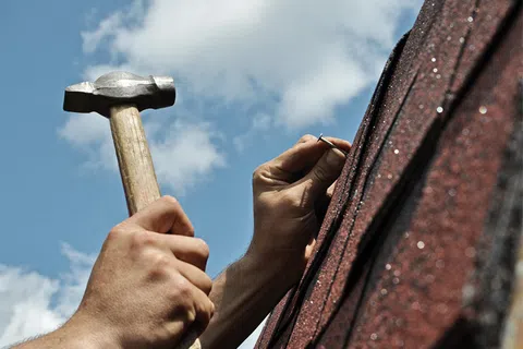 Hands using hammer to nail on textured roofing shingles under a partly cloudy sky outdoors