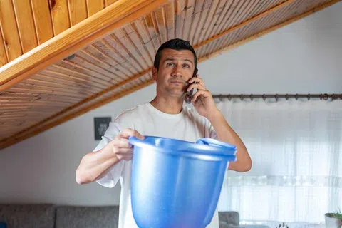Man holding blue bucket under leaking wooden ceiling while talking on phone in living room.