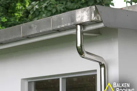 Modern house with shiny stainless steel rain gutter and downspout system on white facade under green trees.