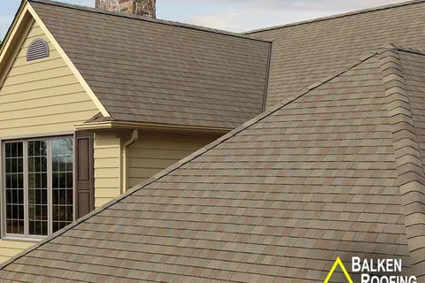 Close-up of residential house roof with brown shingles and stone chimney under a partly cloudy sky.