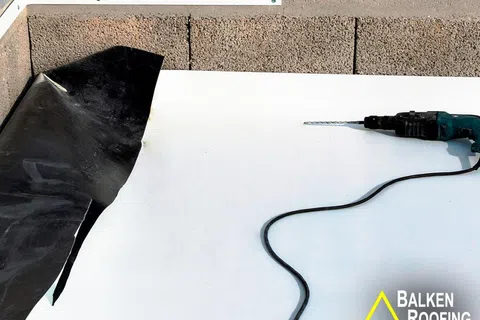 Electric drill resting on white roofing membrane next to black rubber sheet and concrete block wall under construction.