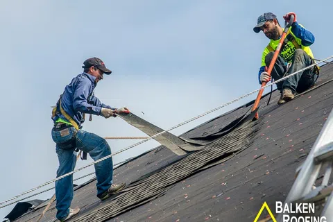 Two workers installing roofing felt on a steep roof using hand tools and safety harnesses under clear sky