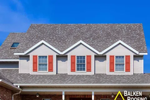 Modern house with gray shingle roofing, beige siding, red window shutters, and clear blue sky background.