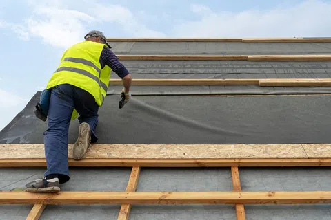 Construction worker in a high-visibility vest installing roofing underlayment on a wooden frame roof structure.