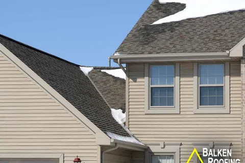 Close-up of house roof with snow patches, beige siding, two windows, and clear blue sky background.