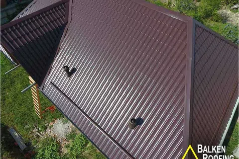 Aerial view of a shiny brown metal roof with vents surrounded by greenery and Balken Roofing logo.