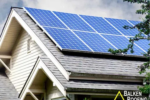 Solar panels installed on a gray shingled house roof under a cloudy sky with green trees nearby