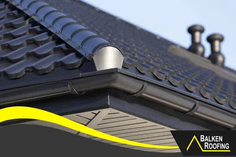 Close-up of black tiled roof corner with gutter system under blue sky and Balken Roofing logo.