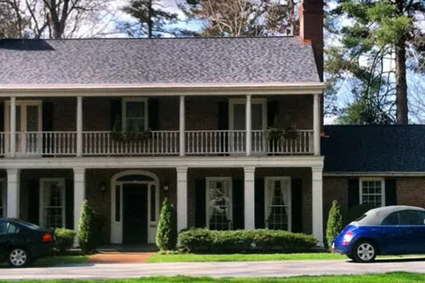 Two-story brick house with white columns, black shutters, balcony, and cars parked in front on a sunny day