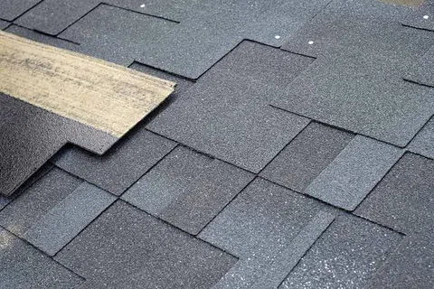 Close-up of dark grey asphalt roof shingles overlapping and textured with one partially lifted shingle.
