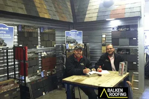 Two men sitting at a table in a roofing materials showroom with shingles displayed on walls