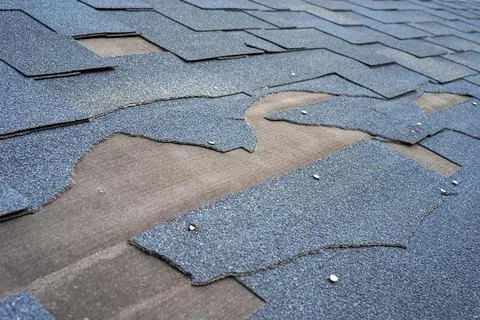 Damaged gray asphalt roof shingles peeling and missing, exposing the wooden roof deck underneath.