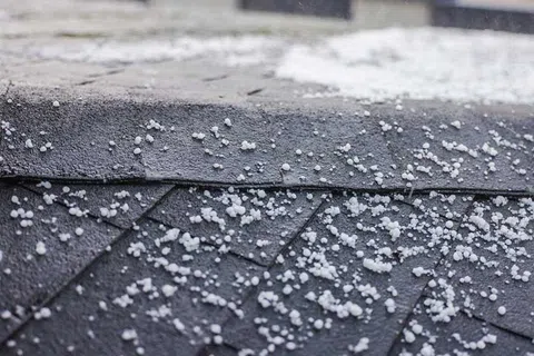 Small hailstones scattered on a dark shingled roof after a hailstorm with droplets of water visible.