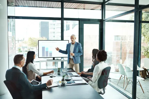 Business team in a modern glass office room during a presentation with charts on a screen.