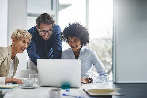Three diverse business professionals gathered around a laptop, smiling and collaborating in a bright office.