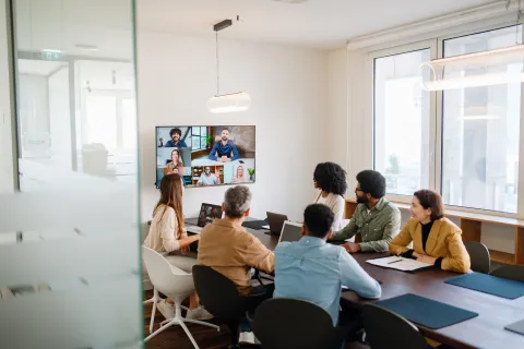Diverse team in office meeting room participating in virtual video conference on wall-mounted screen.