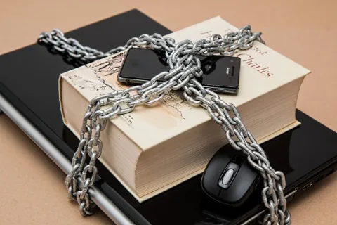 Book, phone, and mouse secured with chained locks on a closed laptop on a brown surface.