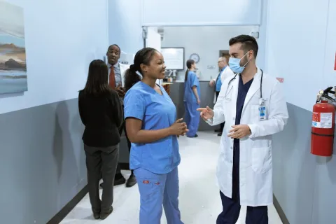 Doctor and nurse wearing scrubs talking in hospital corridor with other medical staff in background.