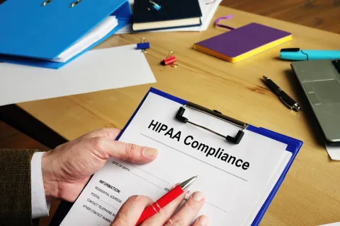 Person holding a clipboard with HIPAA Compliance form on desk with office supplies and laptop.
