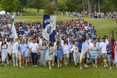 Large group of Berry College students walking outdoors holding flags during an organized event on green lawn.