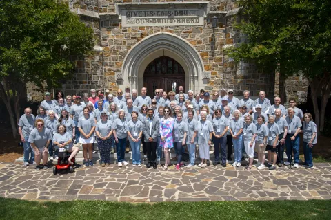 Large group of people in matching gray shirts posing outdoors in front of a stone building with an arched entrance.