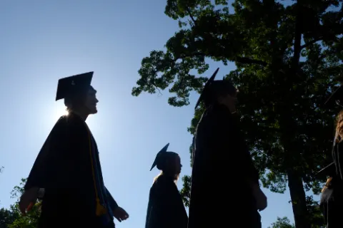 Silhouetted graduates in caps and gowns walking outdoors with trees and clear sky in background at graduation ceremony.