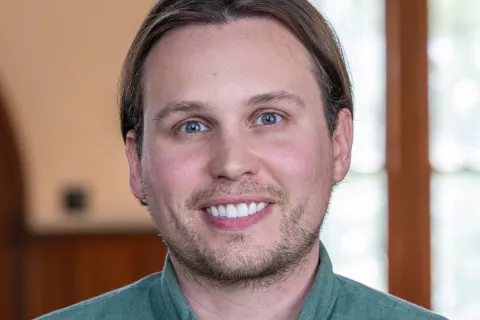 Smiling middle-aged man with beard wearing a blue shirt and black blazer in a softly lit indoor setting