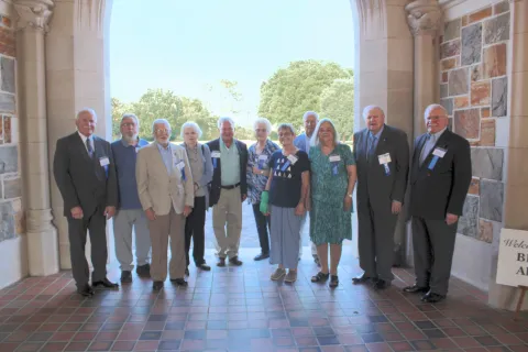 Group of senior adults standing together in a sunlit archway during a formal gathering or reunion.