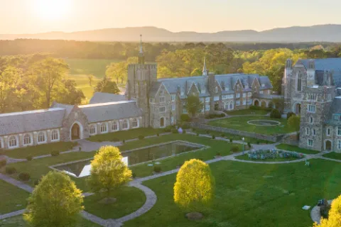 Aerial view of a historic stone campus with green lawns, reflecting pools, and surrounding trees at sunset.