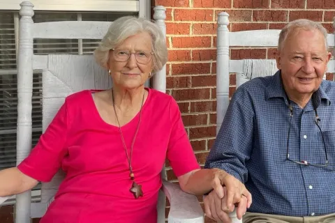Elderly couple holding hands seated on white rocking chairs on a porch with a brick wall background.