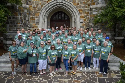 Group of adults wearing matching green Labor Day shirts posing outdoors in front of a stone building.