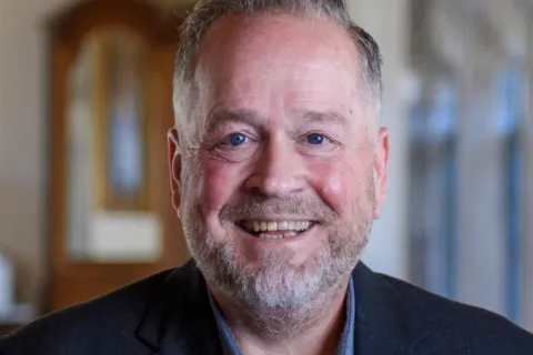 Smiling middle-aged man with beard wearing a blue shirt and black blazer in a softly lit indoor setting