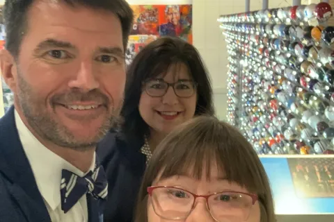 Three people smiling for a selfie in front of a display wall filled with colorful football helmets.