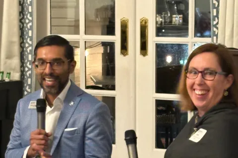 Two people seated indoors holding microphones during a discussion in a cozy room with glass doors behind them