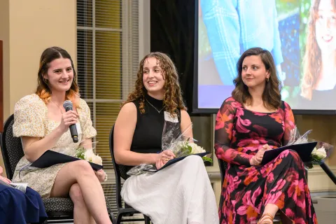 Four women seated indoors at an event holding flowers, one speaking into a microphone with a projected image behind them