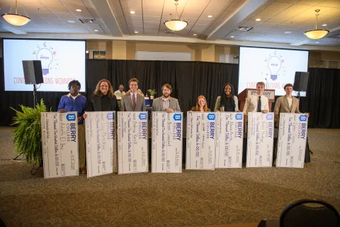 Eight winners hold oversized checks on stage at Berry College competition awards event with light bulb pitch visuals.