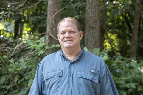 Middle-aged man smiling outdoors wearing a blue short-sleeve button-up shirt with trees and greenery in background