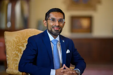 Smiling man in navy suit and glasses seated on ornate chair in softly lit room with blurred background.