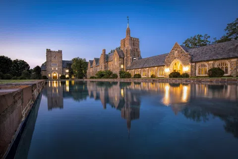 Historic stone building reflected in a calm water pool under a clear twilight sky with warm lights.