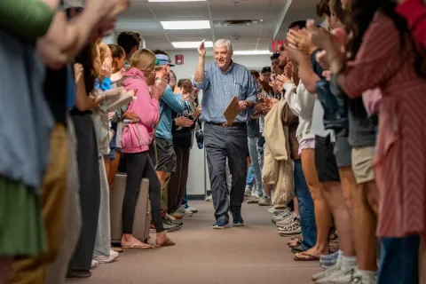 John Grout walking down hallway high-fiving and cheered by people lined up on both sides indoors.